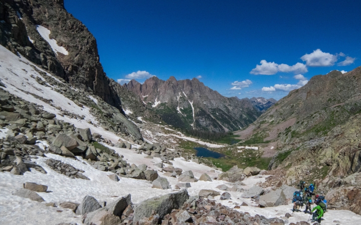 People appear small as they hike in a vast mountainous landscape under a blue sky.